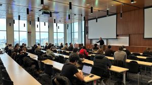A large classroom in Morgridge Hall with floor-to-ceiling windows overlooking campus. Dozens of UW–Madison students sit at long tables listening to Dr. Eric Miller, who is standing at the front of the room speaking during the National Security Career Forum.