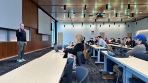 Dr. Eric Miller speaks at the front of a modern classroom as UW–Madison students seated at rows of tables listen and take notes on laptops during the National Security Career Forum.